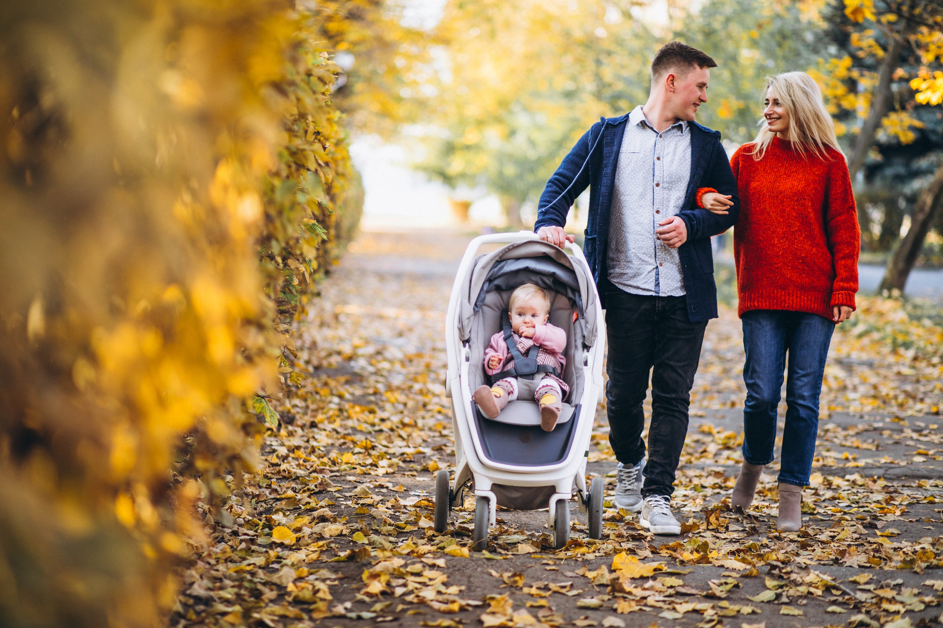 A family enjoying a peaceful walk in the park during autumn, with their baby daughter comfortably seated in a stroller.