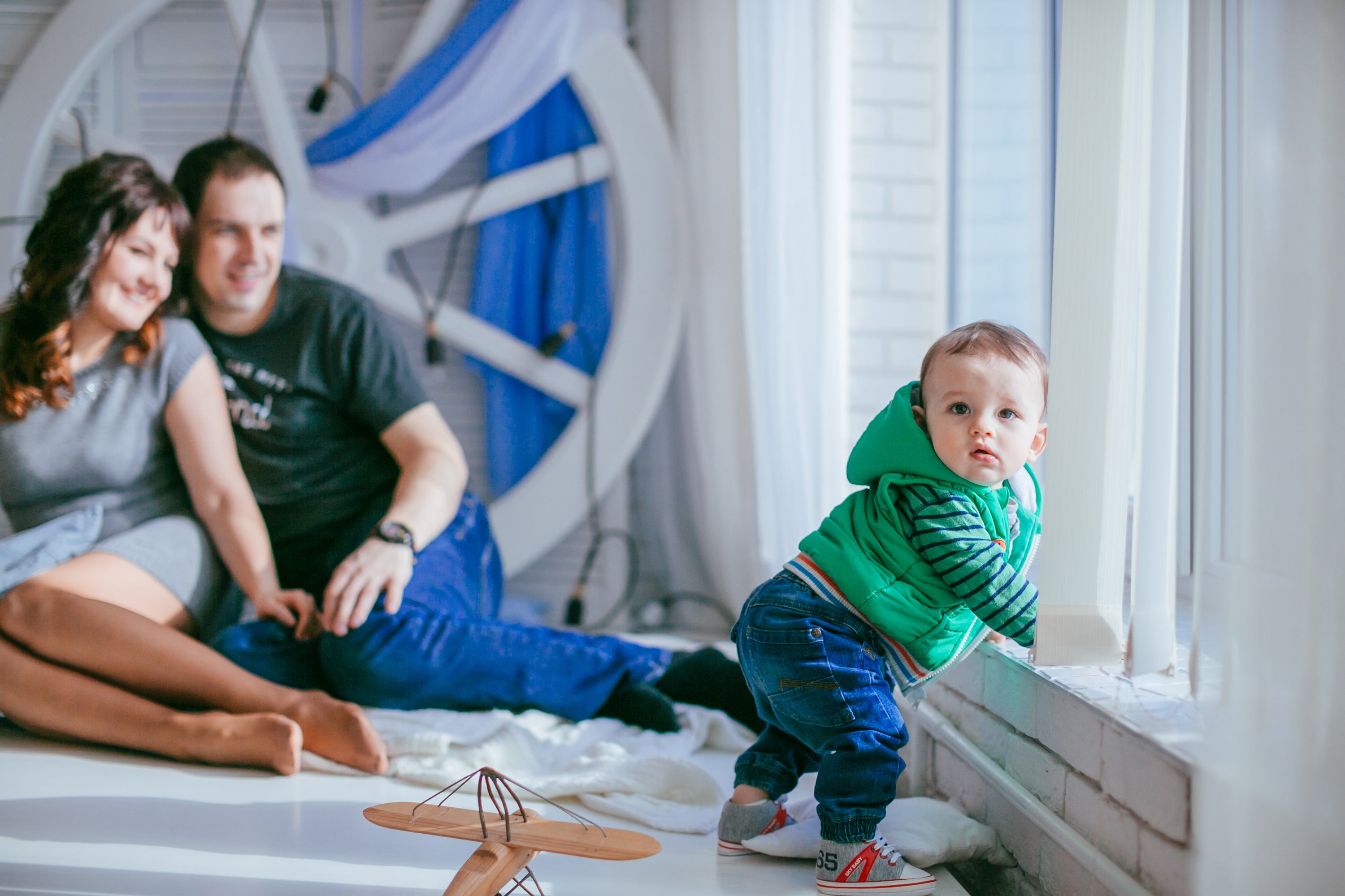 A curious toddler standing by the window while his parents lovingly watch him from the background.