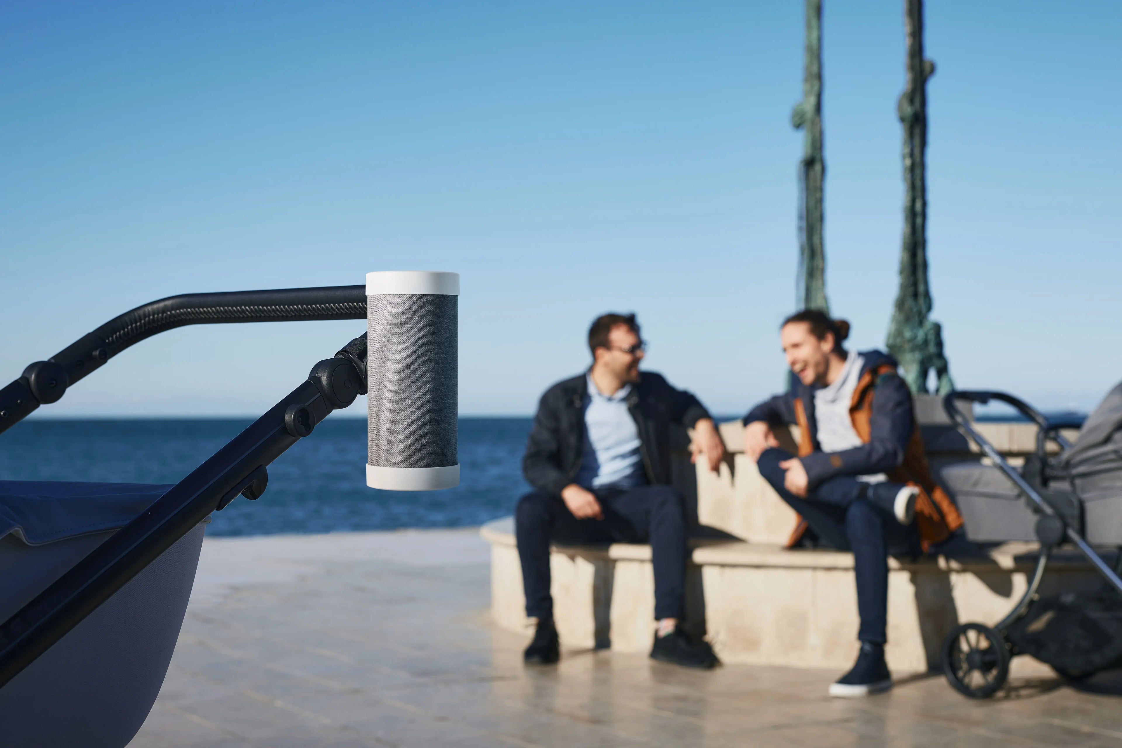 RockingRide® device attached to a stroller handle, with two men sitting and talking by the seaside in the background, showcasing a relaxed outdoor setting.